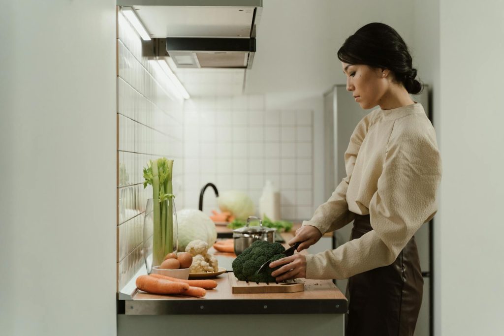 A woman chops broccoli on a kitchen counter with various fresh vegetables around.