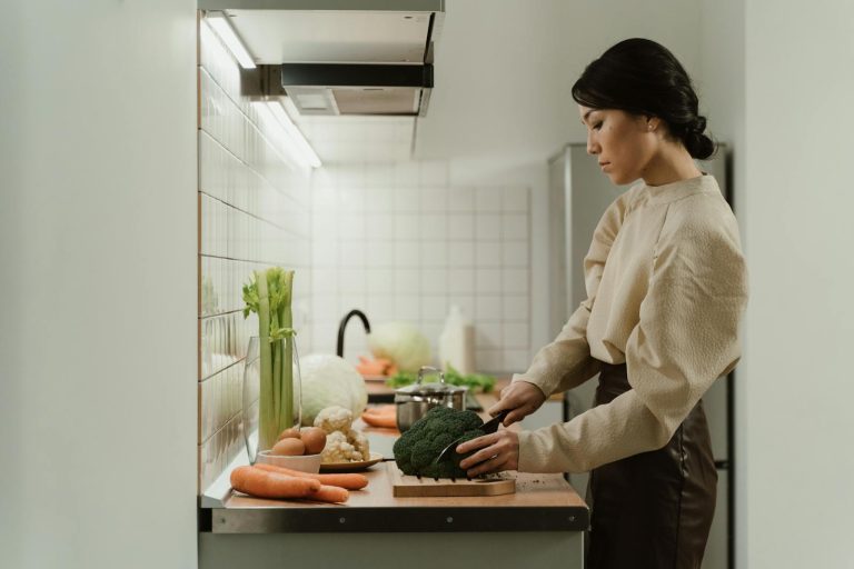 A woman chops broccoli on a kitchen counter with various fresh vegetables around.